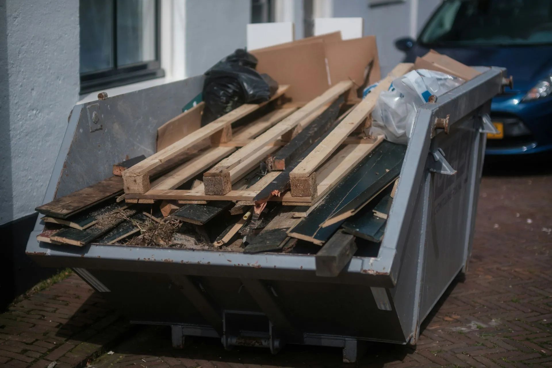 Metal dumpster overflowing with wood scraps and debris; blue car in background.