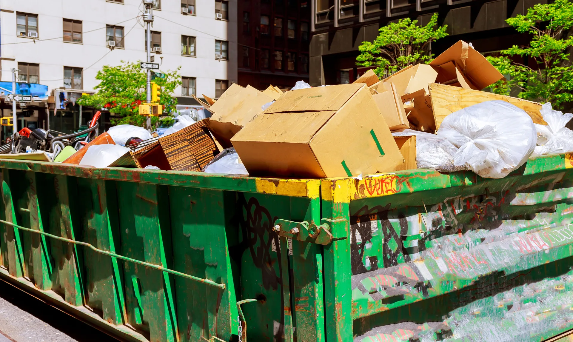 Green dumpster overflowing with cardboard boxes and trash on a city street.