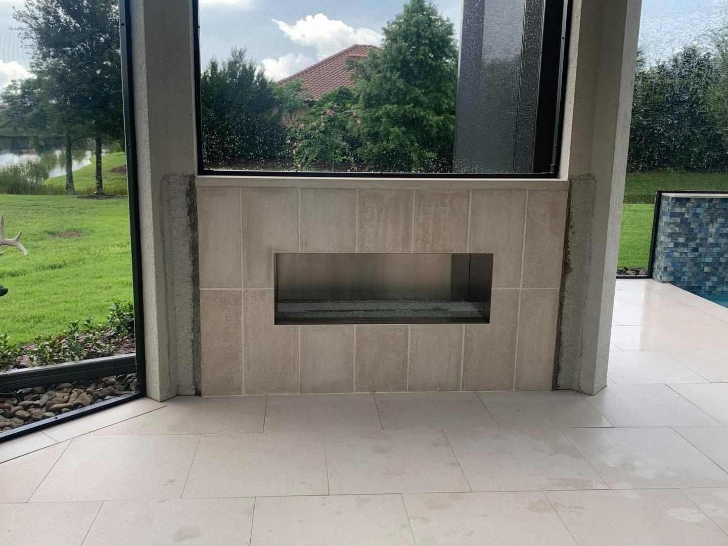Outdoor fireplace with light tile and a built-in niche, framed by glass and overlooking a green lawn.