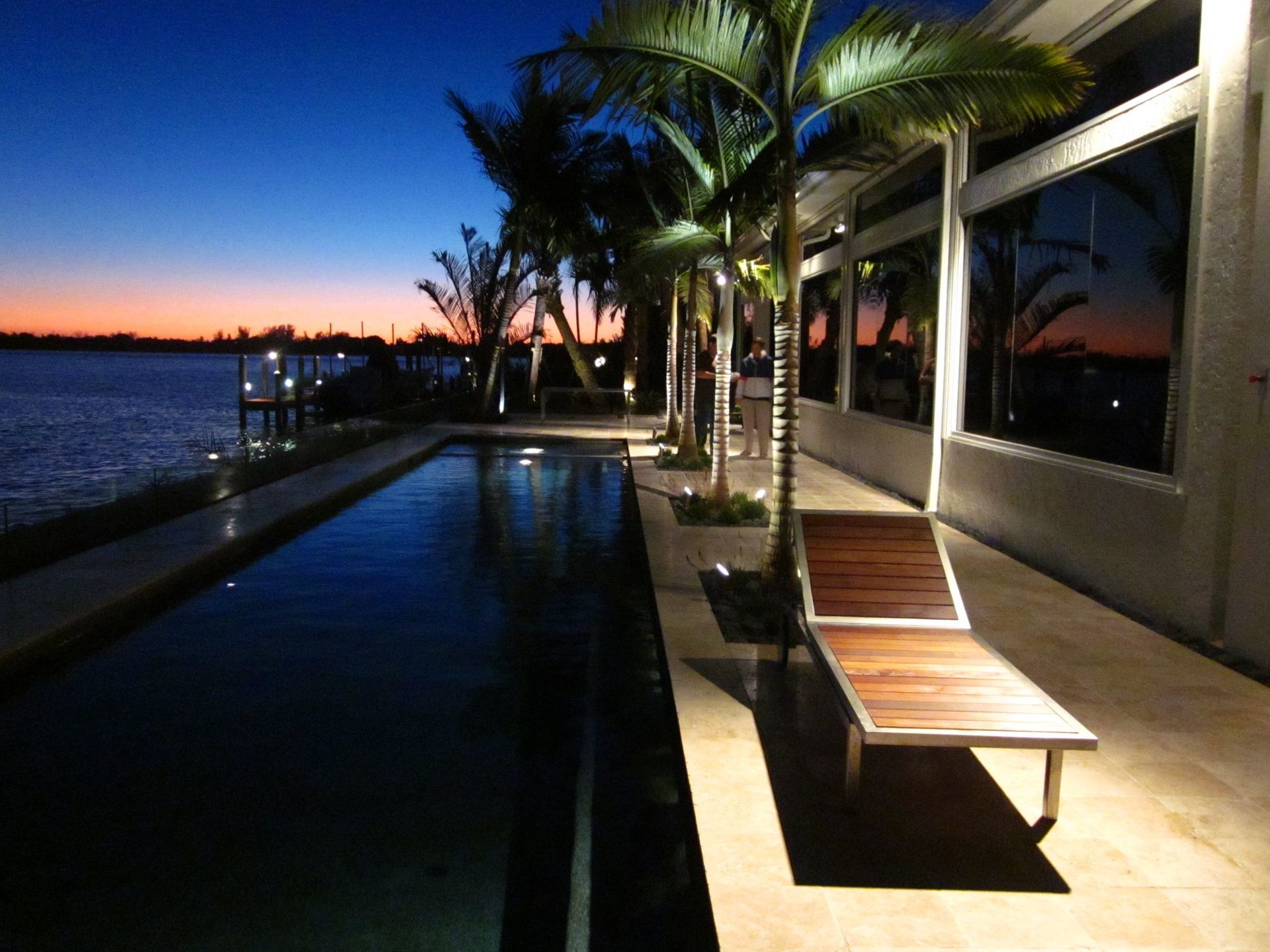 Poolside view at dusk: a lounge chair, palm trees, and a body of water reflecting the colorful sunset.
