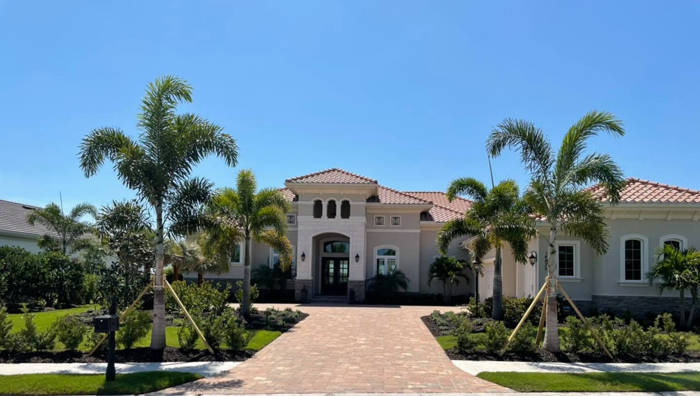 A large, beige house with red tile roof, palm trees, and a brick driveway under a blue sky.