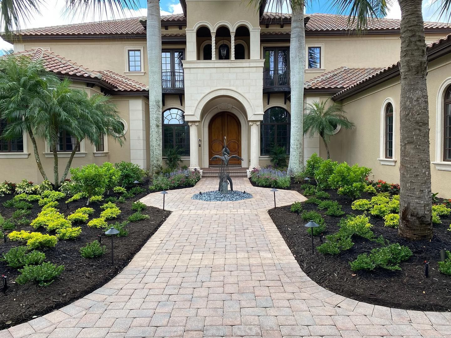 Brick walkway leading to a beige stucco house with arched doorway. Lush landscaping on each side.