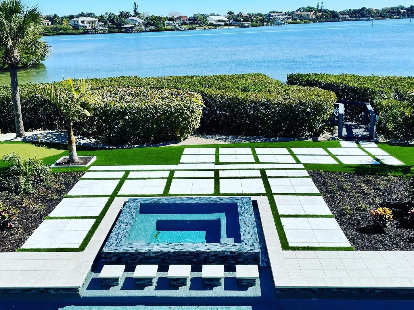 Patio with a pool, water view, greenery, and stone walkway on a sunny day.