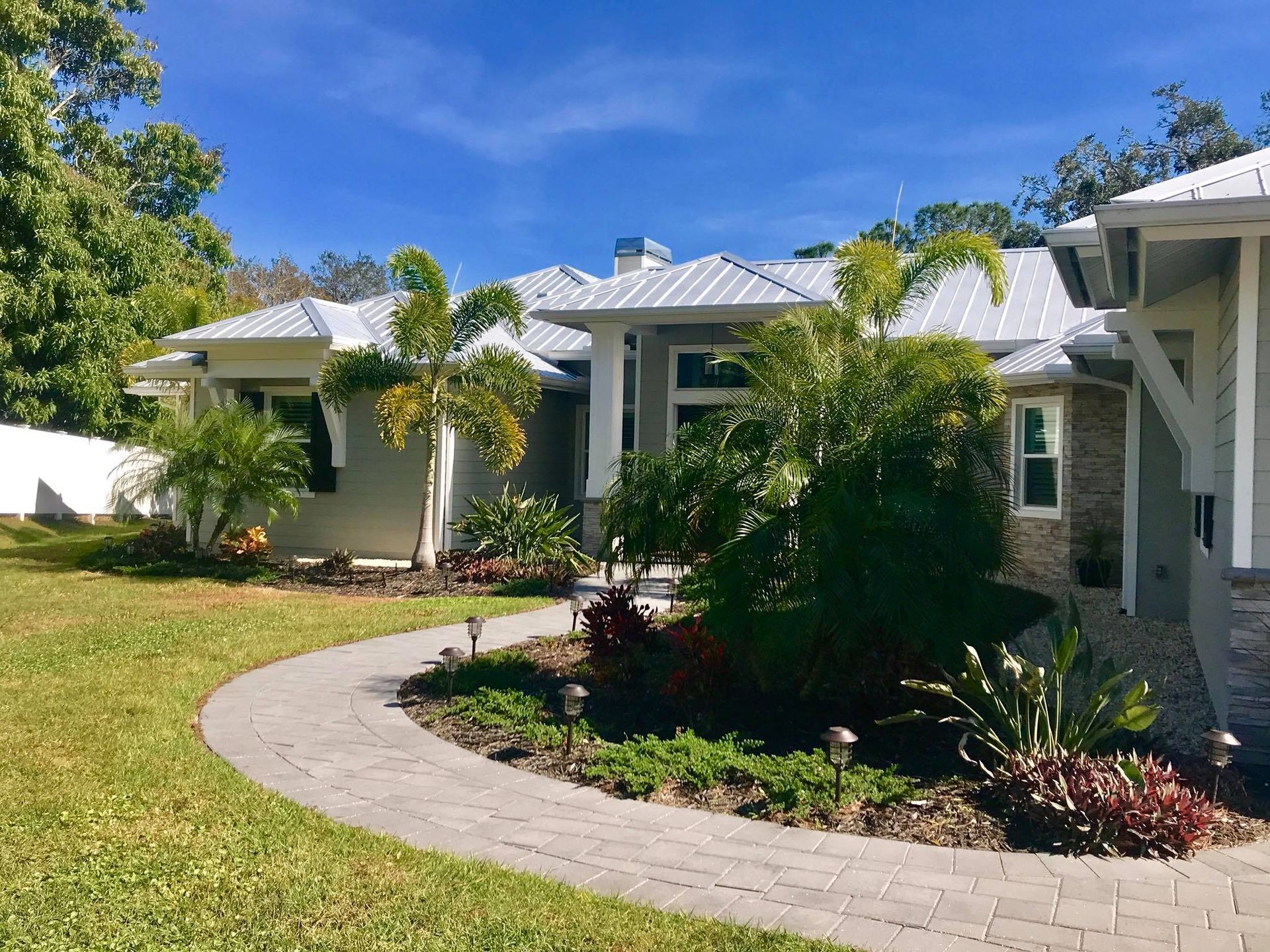 House with gray siding, brick pathway, and tropical landscaping under a blue sky.