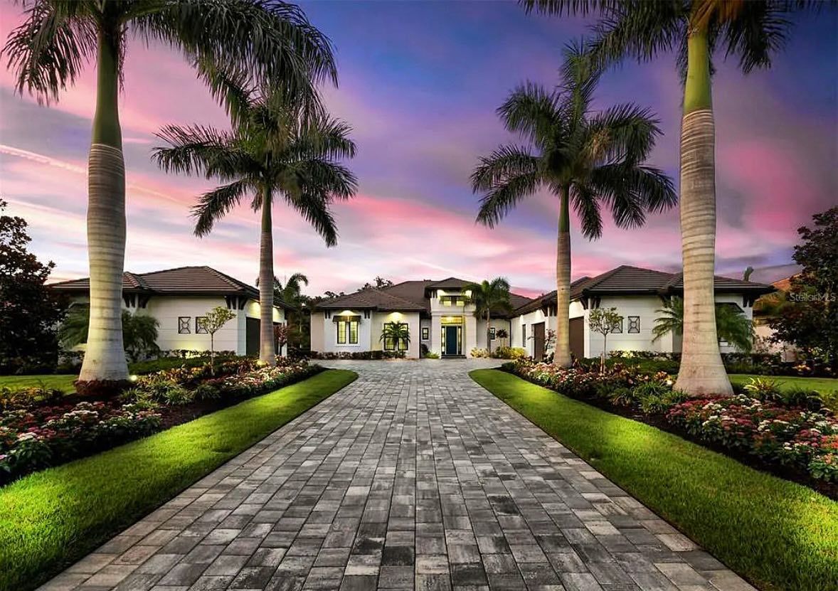 A grand house with a brick driveway lined with palm trees and illuminated hedges, against a sunset sky.