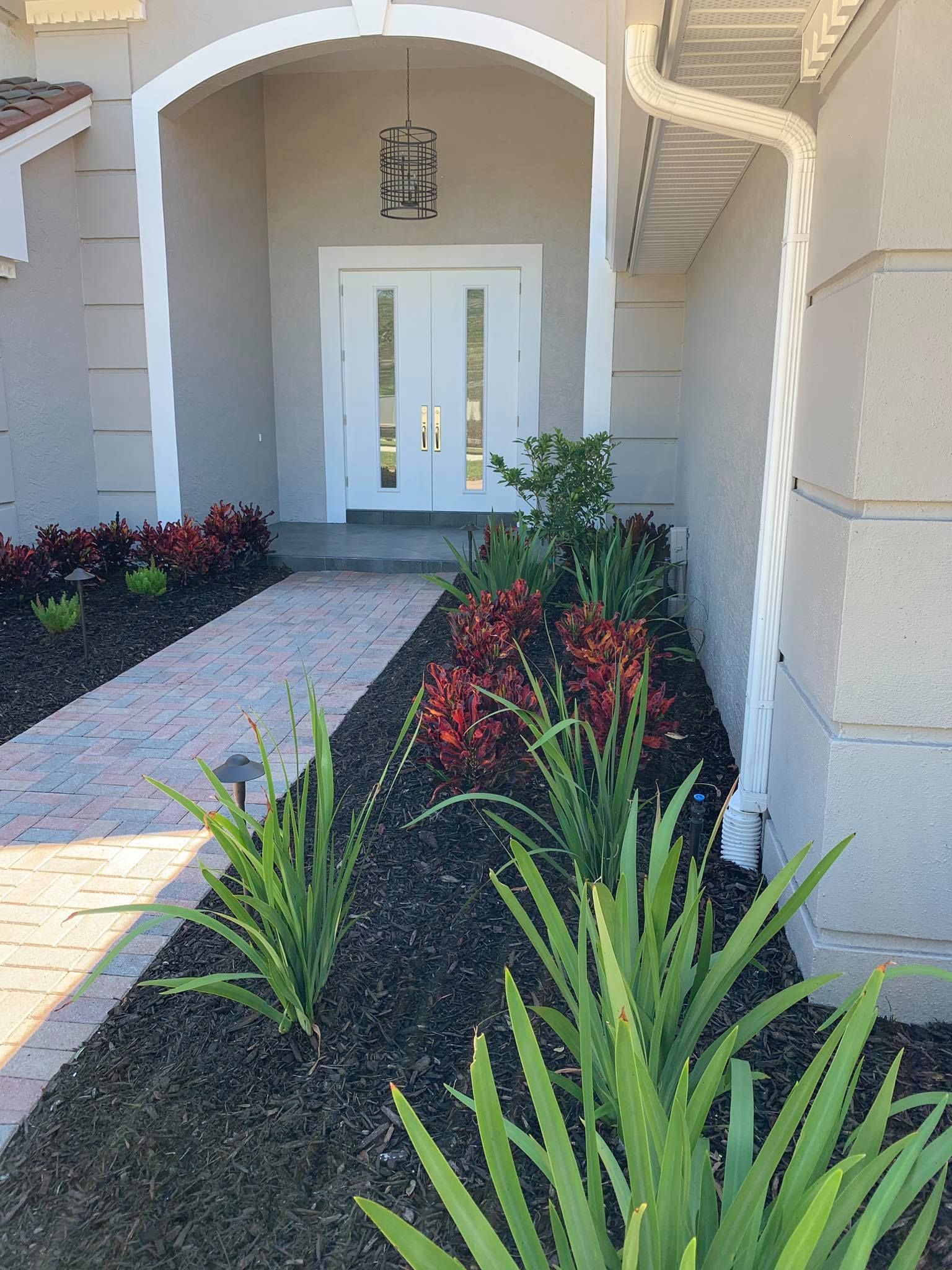 A brick pathway leads to a white double-door entry with a landscaped bed of plants in front.