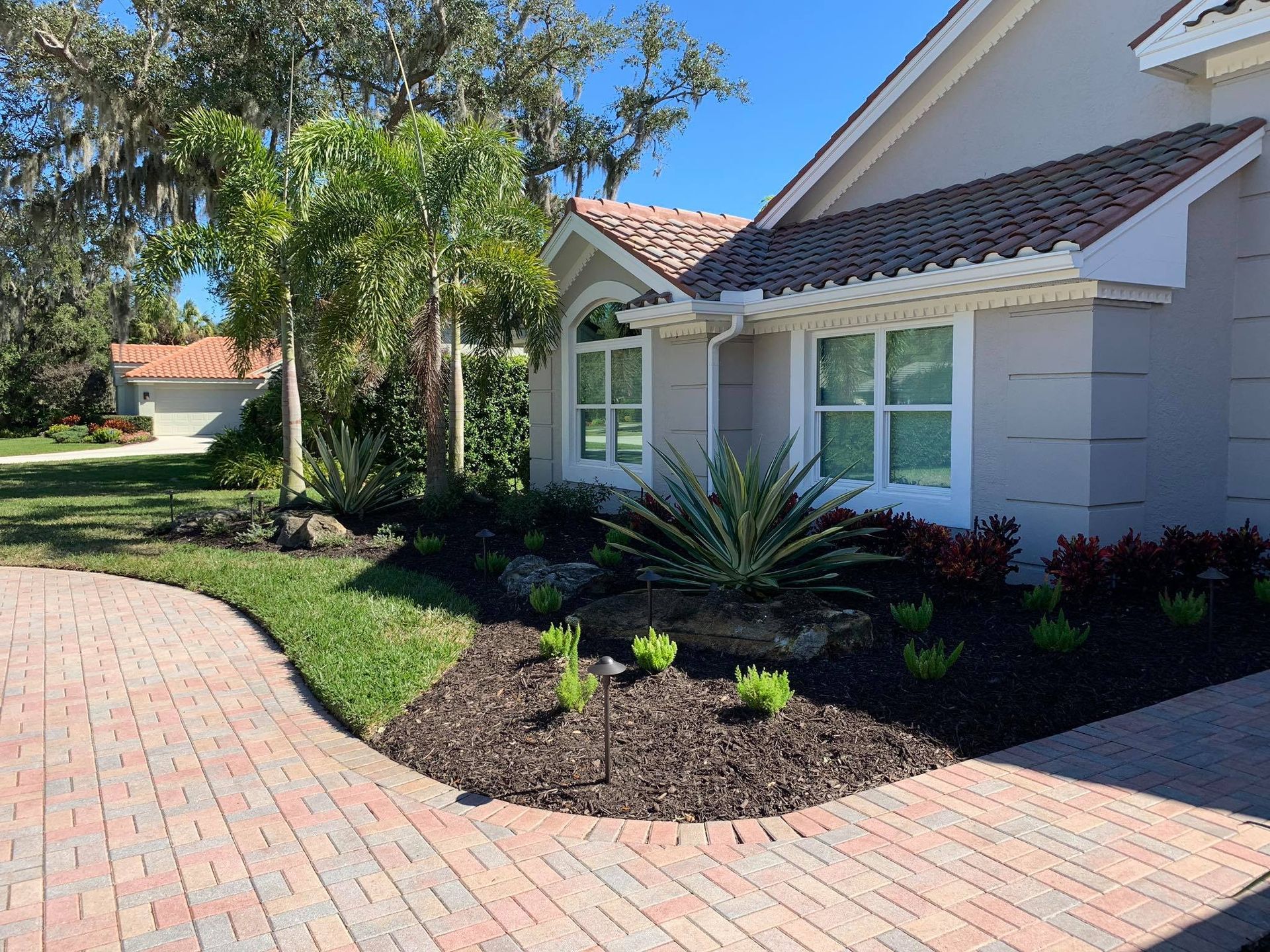Brick pathway leading to a light gray house with red tile roof and tropical landscaping.