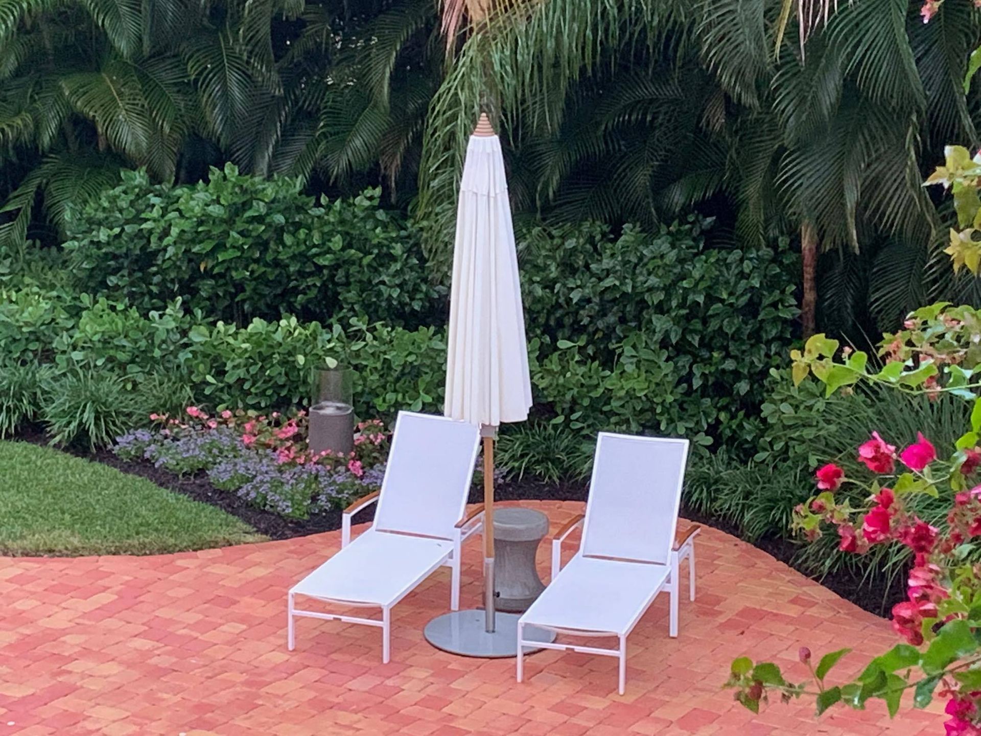 Two white chaise lounges under a closed cream umbrella on a brick patio, surrounded by greenery.