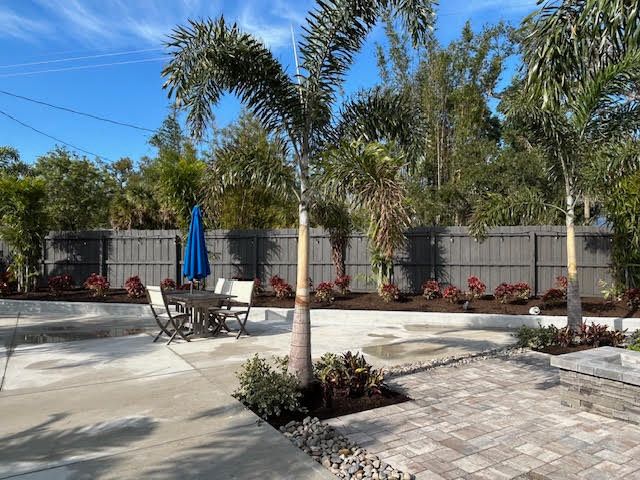 Backyard patio with palm trees, dining set, and fence. Sunny day with blue umbrella.