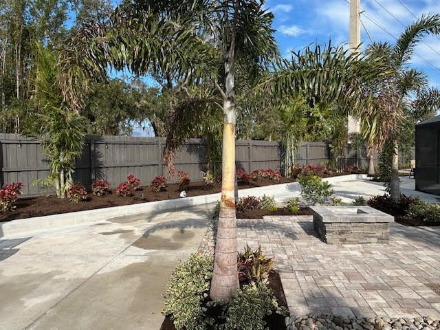 Backyard landscaping with palm trees, dark fence, paved patio, and colorful shrubs.