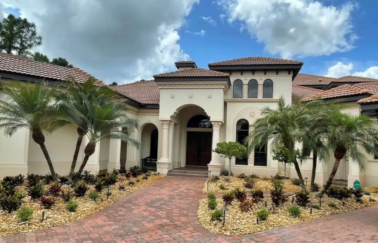 Beige luxury home with red brick pathway, palm trees, and manicured landscaping under cloudy sky.
