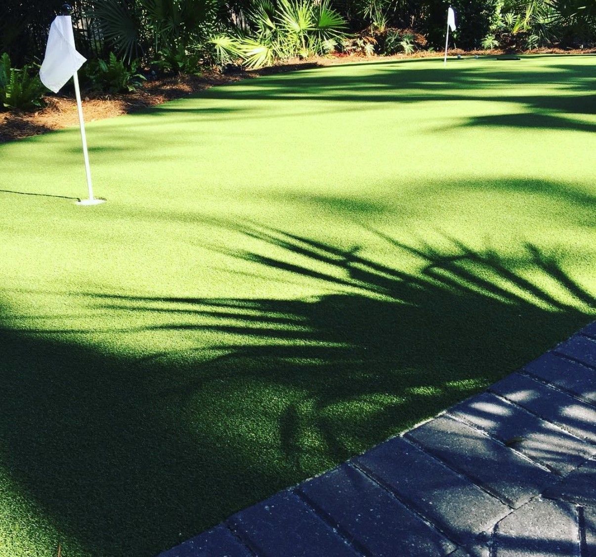 Green artificial putting green with white flags and dark shadows, near a brick border.