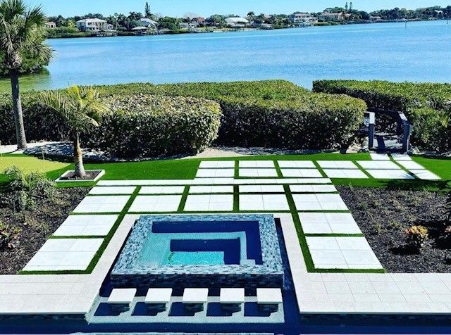 A luxurious outdoor patio with a pool overlooking a lake. Features a modern design with grass and square pavers.