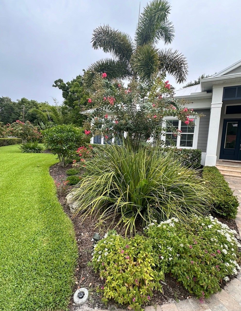 Landscaped front yard with lush green grass and colorful plants bordering a home under overcast skies.