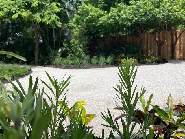 Gravel path through lush green foliage, leading towards trees and a wooden fence.