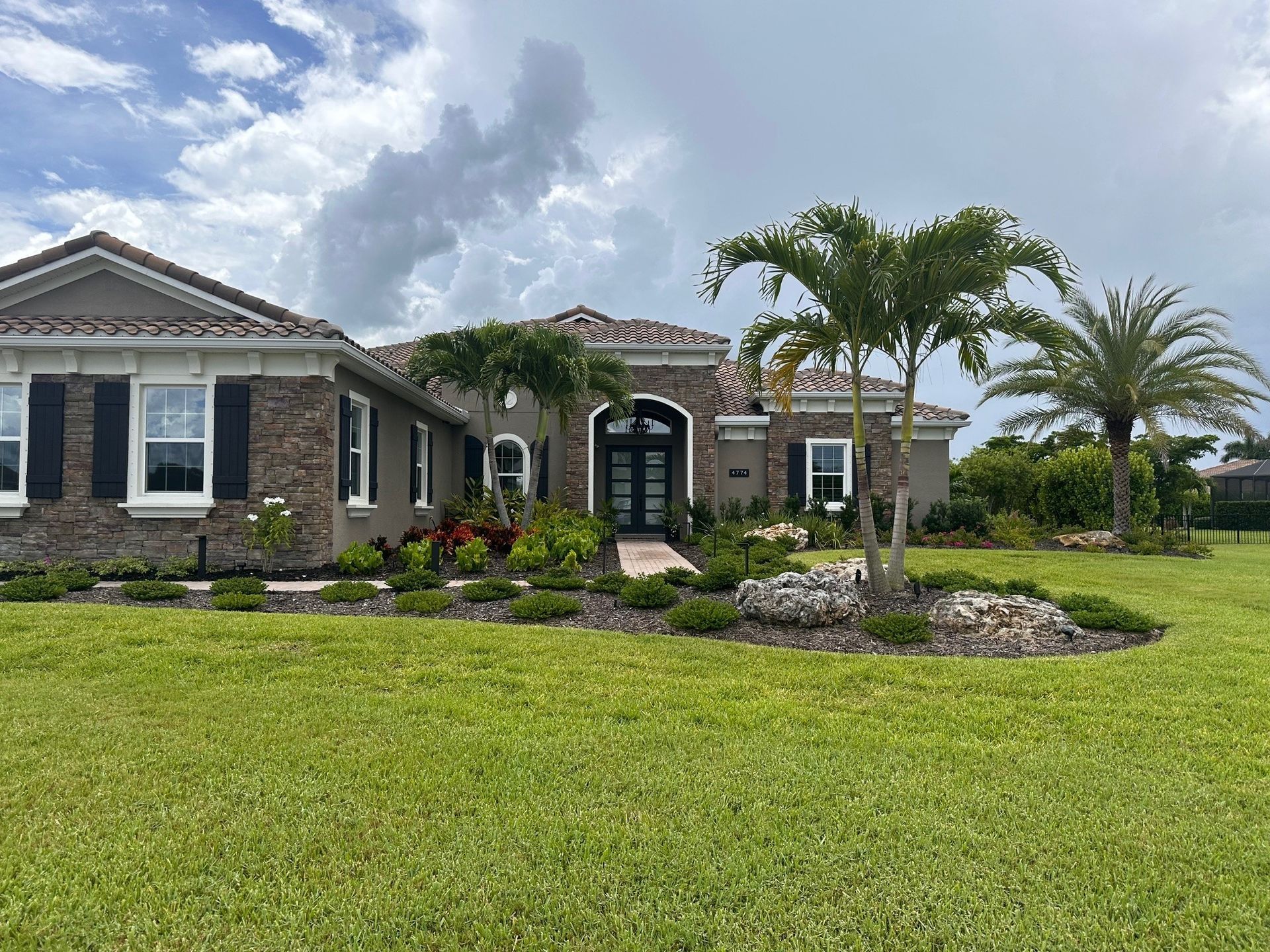 A single-story house with lush landscaping. The home has a red tile roof, brick and stucco exterior, and palm trees in front.