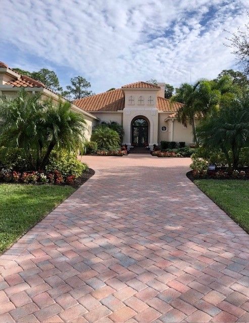 Paver driveway leads to a beige house with a tile roof, surrounded by lush landscaping. Blue sky with clouds.