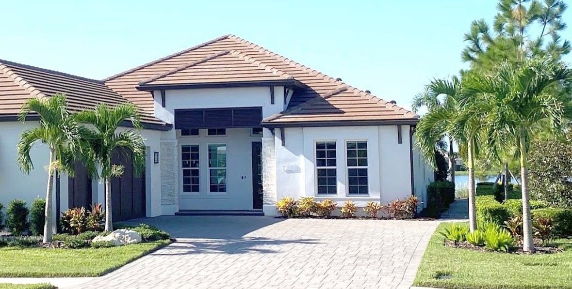 House with white exterior, brown tiled roof, palm trees, and a brick driveway.