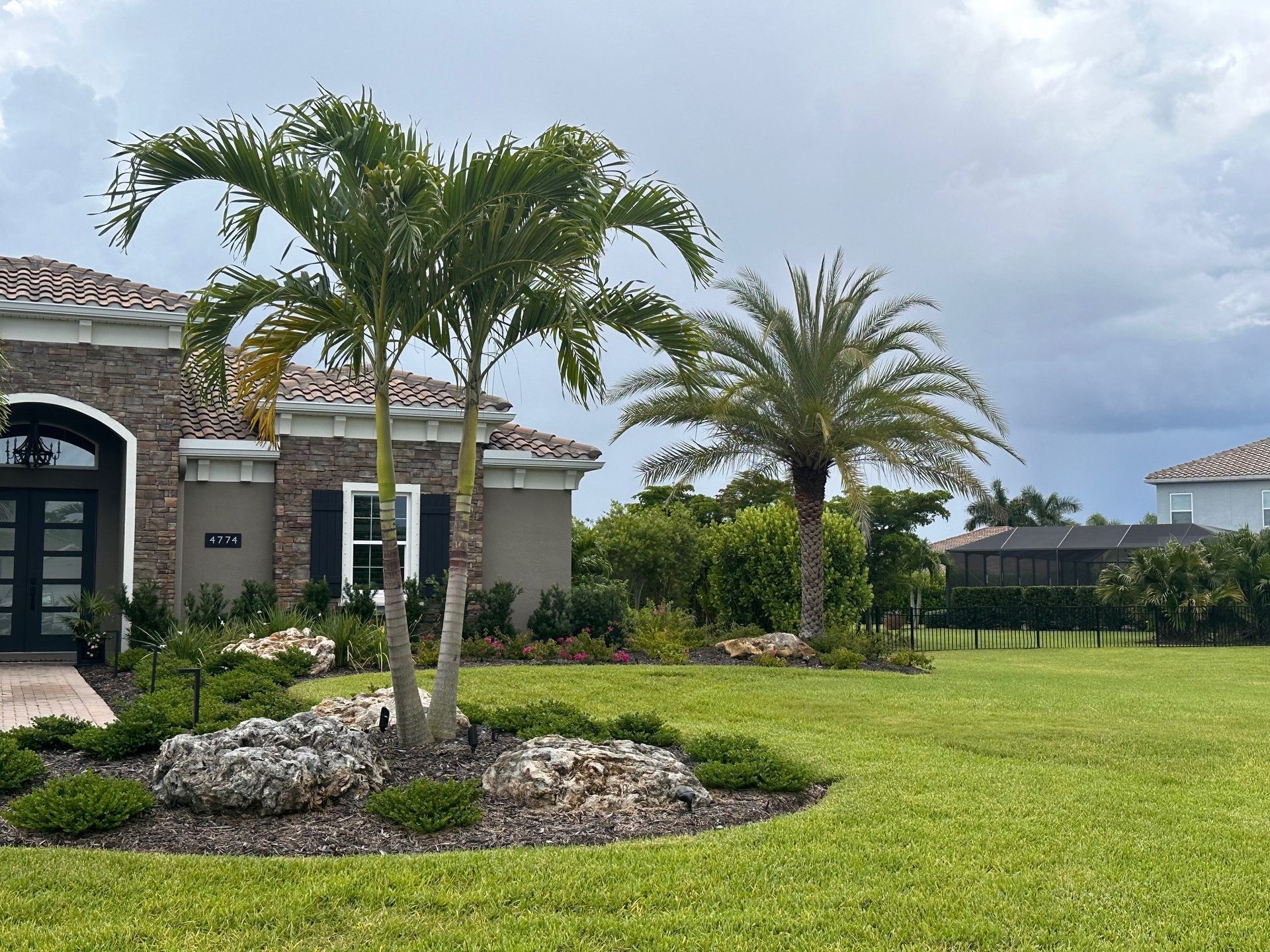 Palm trees and landscaping in front of a house with a gray stucco exterior and a brick facade.