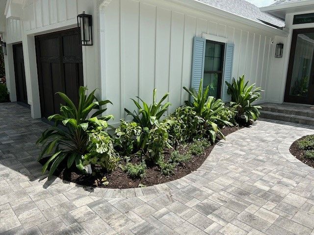 Landscaped flower bed with green plants next to a white house and brick walkway.
