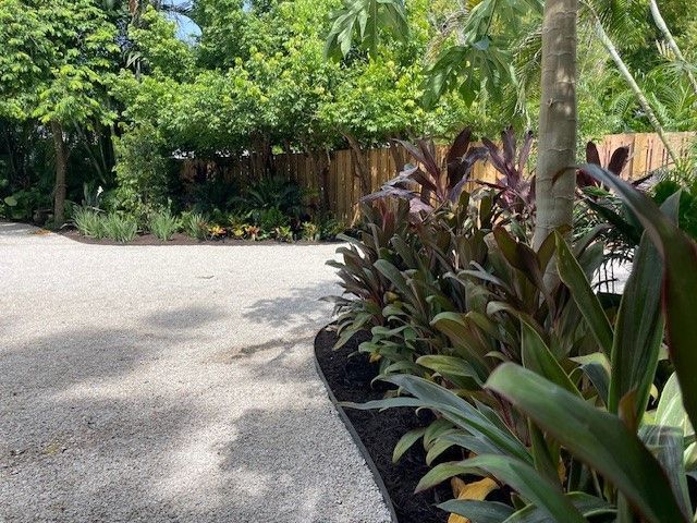 Gravel driveway curves past lush tropical plants with red and green leaves, sunny outdoor setting.