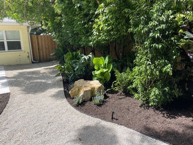 Gravel path curves past a landscaped garden with lush greenery, mulch, and a large light-colored stone.