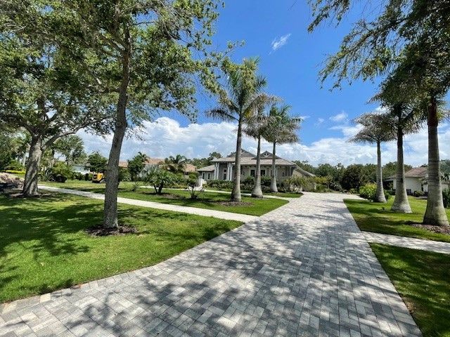 Stone driveway leading to a large house lined with palm trees and grass under a blue sky.