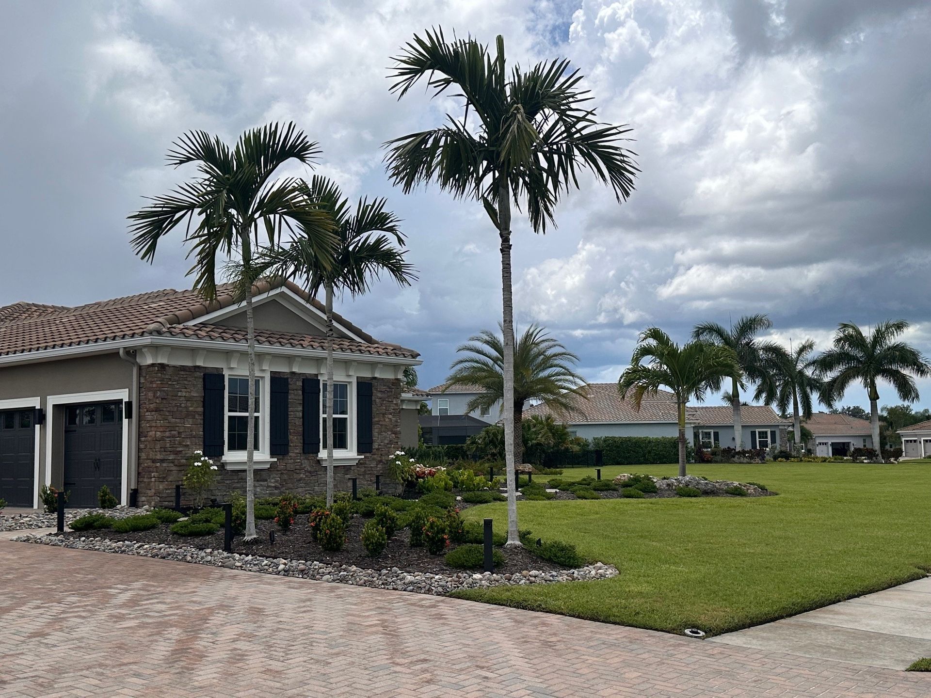 Residential area with palm trees, brick driveway, and houses with green lawns under a cloudy sky.