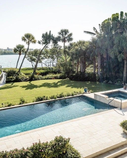 A long, blue pool and spa next to a green lawn, with a water view and palm trees.