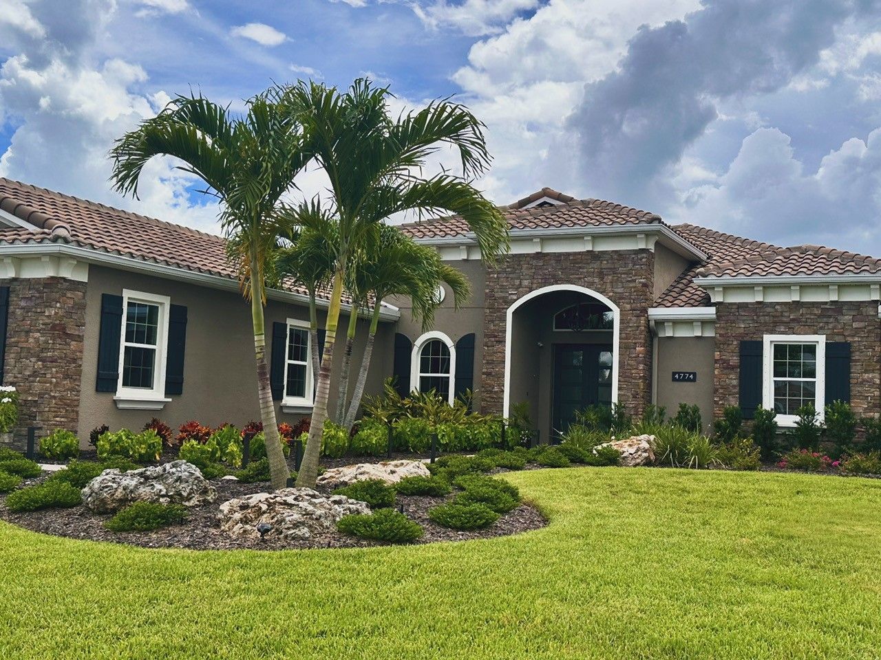House with stucco and stone facade, red tile roof, palm tree in front, green lawn, cloudy sky.