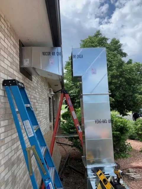 HVAC ductwork installation on a brick building exterior, with ladders leaning against the wall.