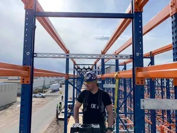 Man in hard hat on scaffolding, surrounded by blue and orange industrial shelving.