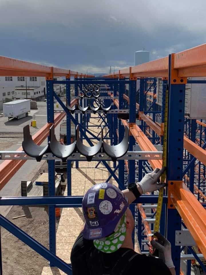 Worker measuring blue and orange industrial shelving, with a tape measure outdoors under a cloudy sky.