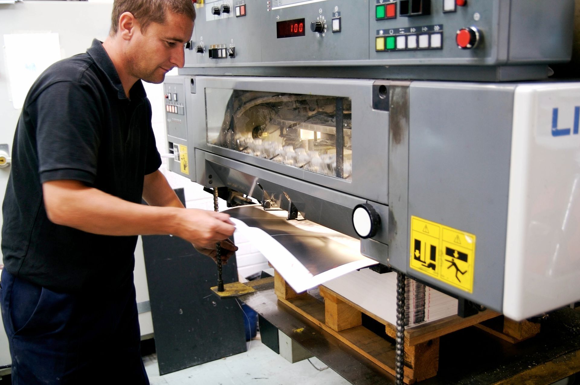 Man operating a large paper cutter machine in a printing facility, focused on the paper.