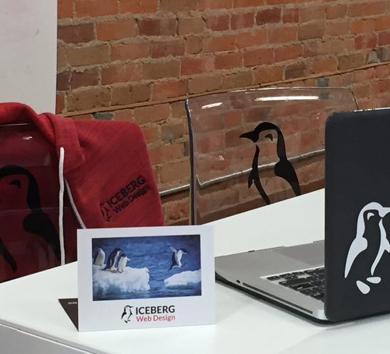 Desk with a laptop and penguin-themed items, including a card, hoodie, and chair, in a brick-walled office.
