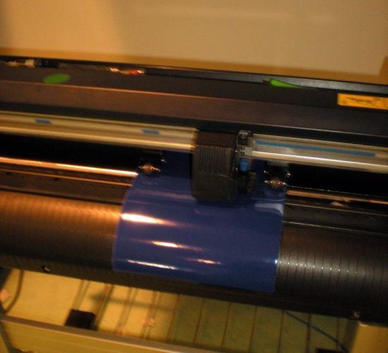 A vinyl cutter with blue vinyl loaded. Black and silver machine, close-up shot.