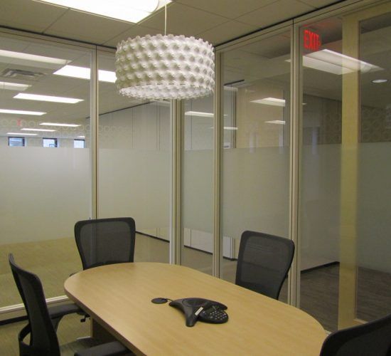 Conference room with oval table, black chairs, frosted glass walls, and a decorative pendant light.