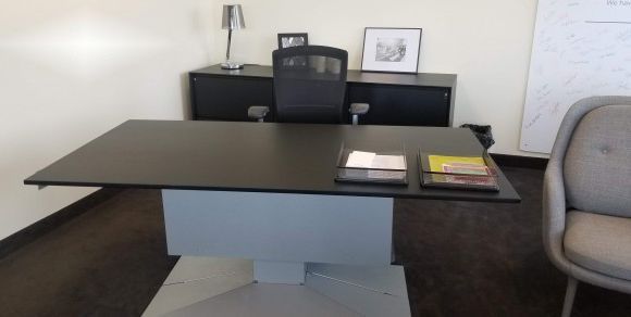 A modern office desk with a black surface and a white pedestal base. A black office chair, a side cabinet, and a gray armchair are also visible.