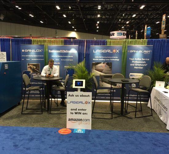 A trade show booth with signs, a person, tables, chairs, and a giveaway sign.