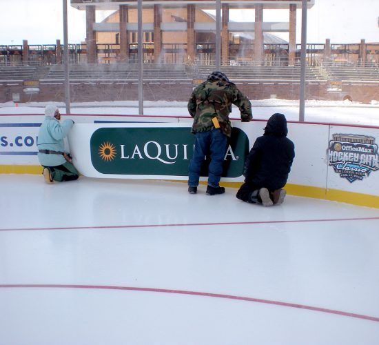 Three people installing a sign on an outdoor hockey rink. Sunny day, snow on the ground, La Quinta logo visible.