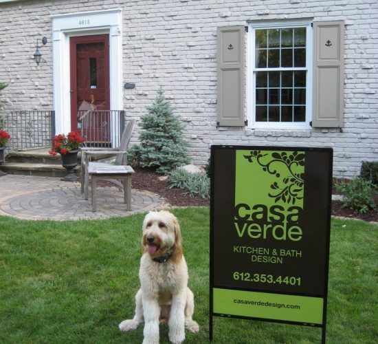 A golden doodle sits in front of a Casa Verde Kitchen & Bath Design sign in front of a house.