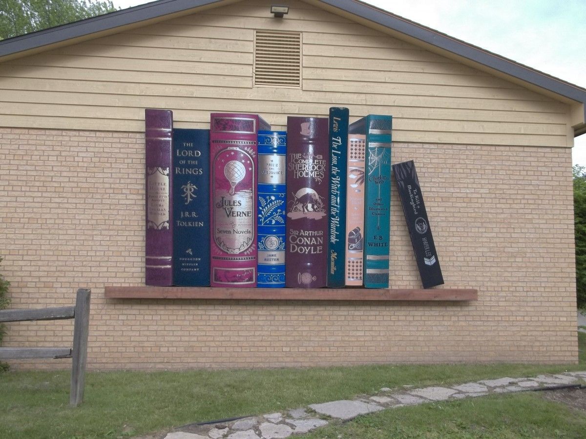 Mural of large books on a brick wall, depicting a bookshelf; purple, blue, and gold titles.