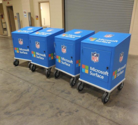 Four blue Microsoft Surface charging stations on wheels, branded with NFL logos, on a concrete floor.