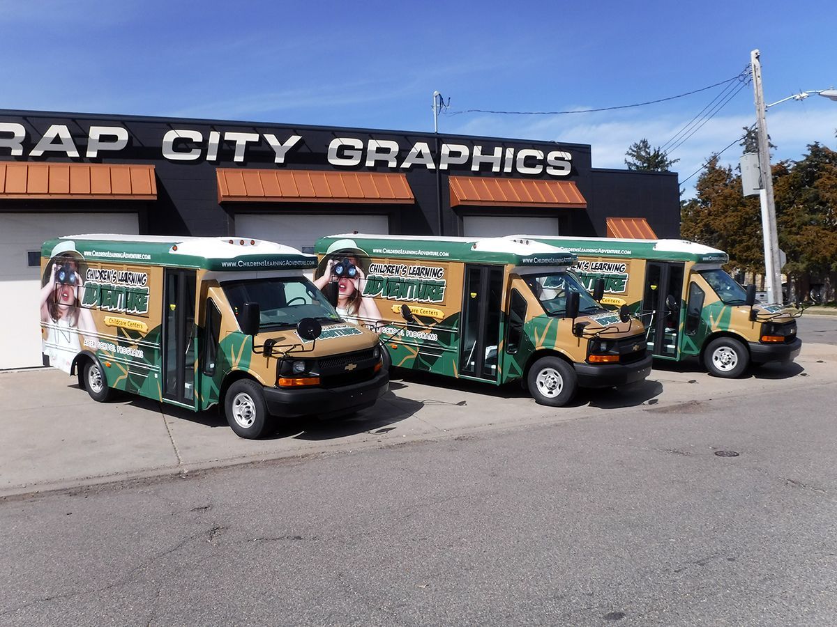 Three wrapped buses in front of "Wrap City Graphics" building. Green, tan camouflage wrap. Sunny day.