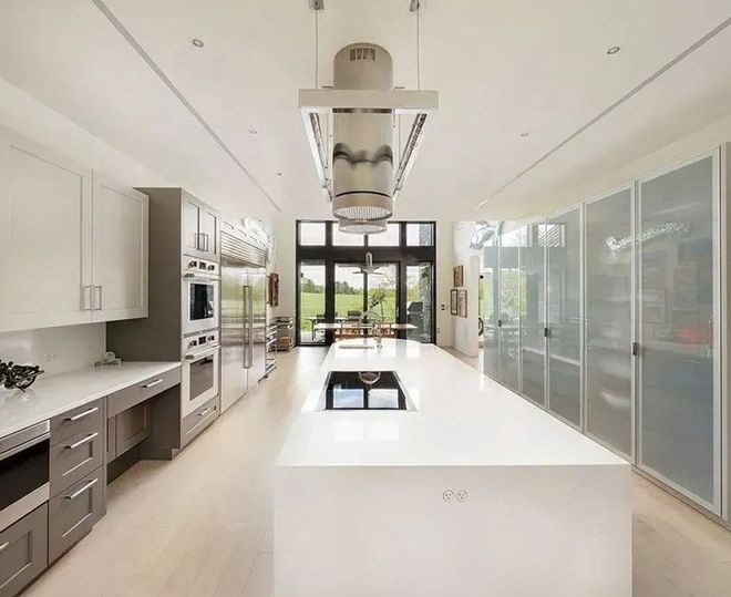 Modern white kitchen with large island, stainless steel appliances, and glass doors to a backyard view.