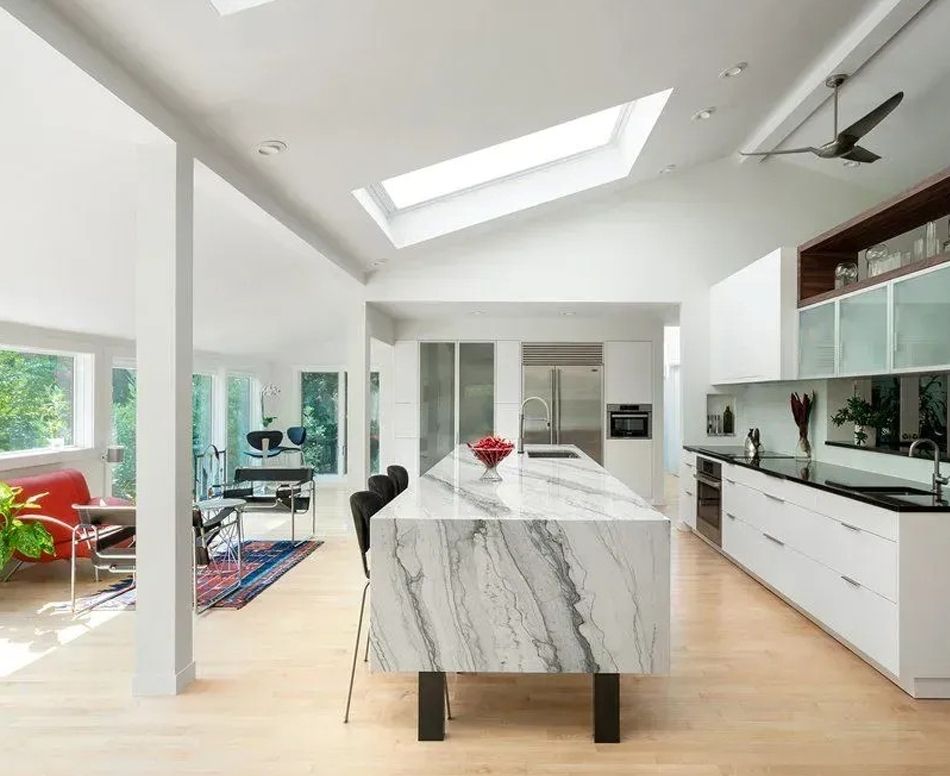 Modern kitchen with large marble island, white cabinets, skylight, and open to living space with windows.