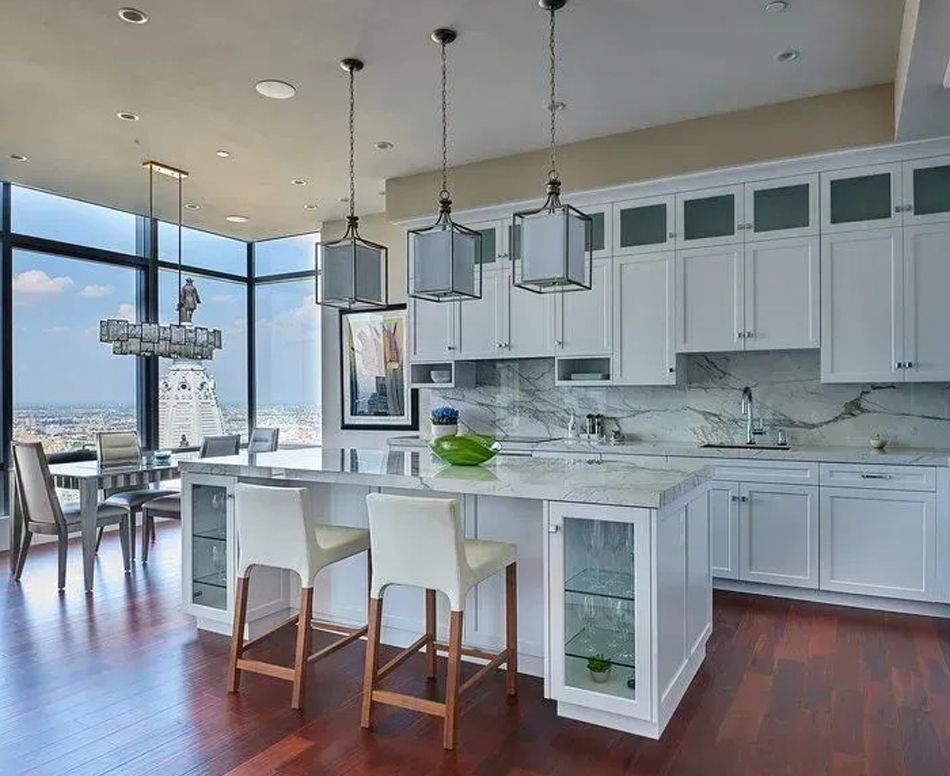 Modern white kitchen with island, seating, and city view. Dark wood floors.