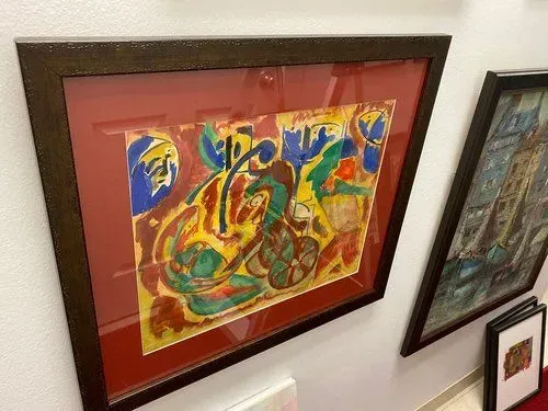 Abstract artwork in a brown frame with red matting. Colorful shapes and a blue face on a white wall.