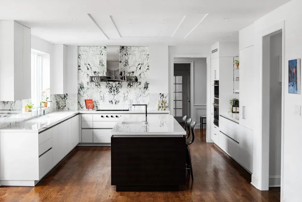 Modern white kitchen with dark wood island, marble backsplash, stainless steel appliances, and wood floors.