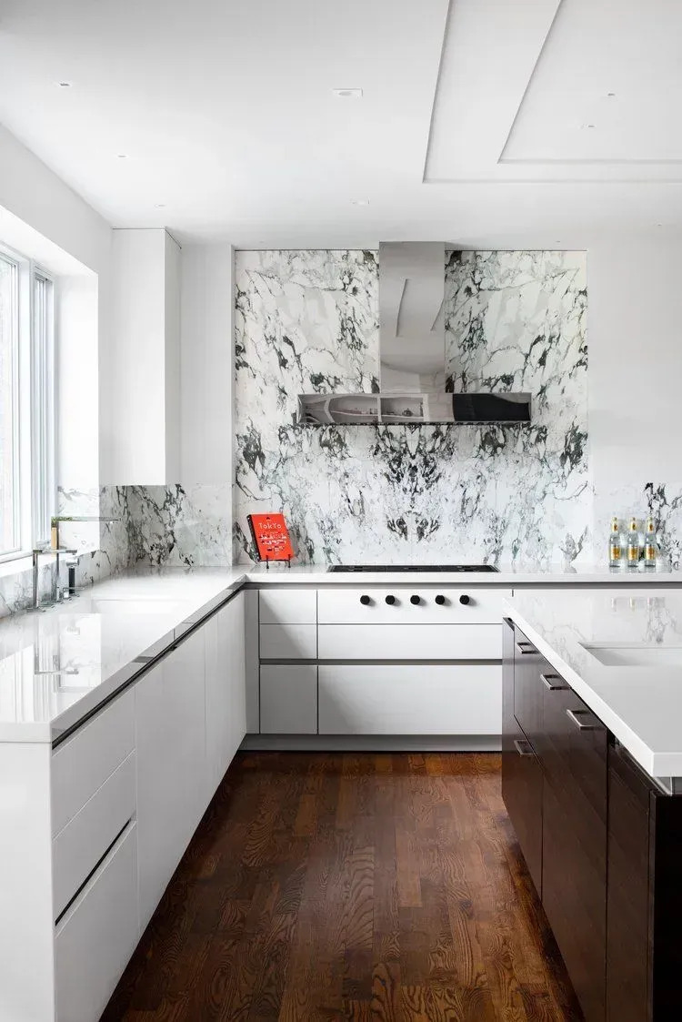 Modern white kitchen with dark wood floor, marble backsplash and countertops, and stainless steel appliances.
